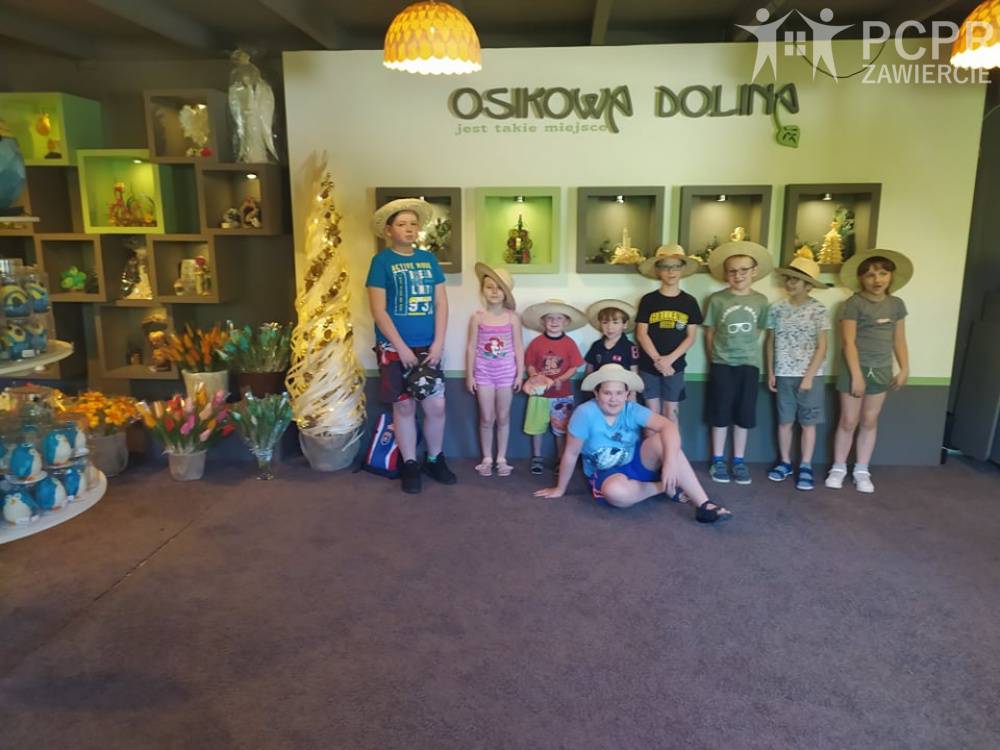Zdjęcie: Grupa dziewczynek i chłopców w kapeluszach na tle wyrobów z osikowego drewna